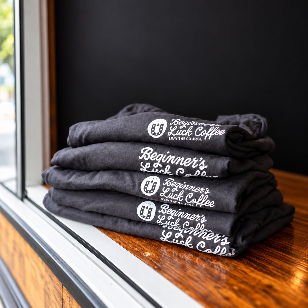 A stack of folded Beginner's Luck Coffee shirts on a windowsill